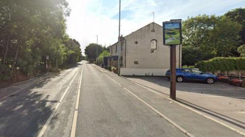 The A58, which runs past the Valley Inn pub. A blue car is parked next to the pub.