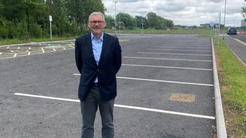 A man standing in an empty car park. He is wearing a suit jacket and shirt and has his arms behind his back.