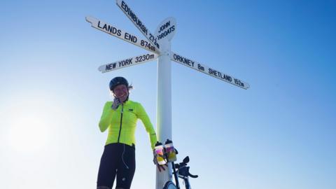 A woman standing in front of the Land's End sign with a bicycle