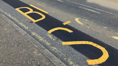 A bus stop road marking that has only been partly repainted over. The b and s in the words bus stop are clear whereas the rest of the words have worn away.