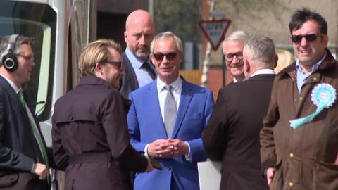 Nigel Farage is standing in the street surrounded by several people. He is wearing a blue suit, white shirt and grey tie.