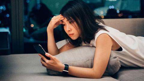 A woman lies on her front on a lounge, holding a phone in one hand and pressing her brow with the other