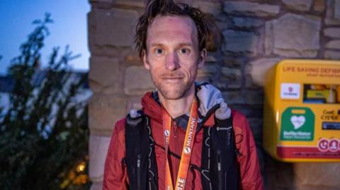 A man with dark hair is looking into the camera with a slight smile. He is wearing a red coat and an orange lanyard, and black rucksack straps are over his shoulders. Behind him is a yellow defibrillator box.