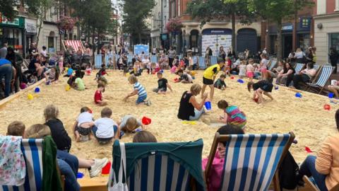 Children and adults play in the sand at the beach. Buckets and spades can be seen as well as rows of deck chairs. In the background are a bunch of cafes and shops as well as trees.