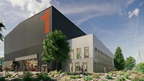 A tall dark grey block with 1 painted on its right side in orange. A two-storey light grey building with windows sits next to it. There are trees and plants with Sunderland's Northern Spire bridge in the background. 