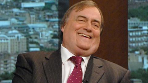 A smiling John Prescott appearing on TV wearing a brown pin-striped suit and red tie. 