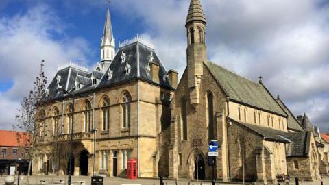 Bishop Auckland's Town Hall and St Anne's Church. The stone buildings are next to each other with a small square and red telephone box in front of them.