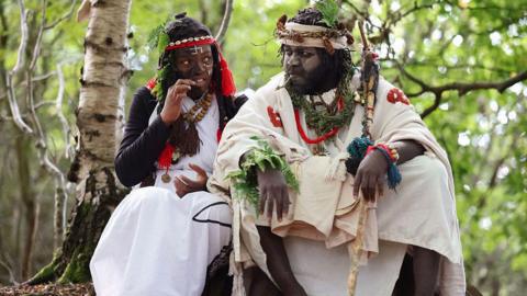 A man and a woman dressed in white/cream robes in a woodland setting. They have painted faces and head-dresses. They have leaves in their hair. 