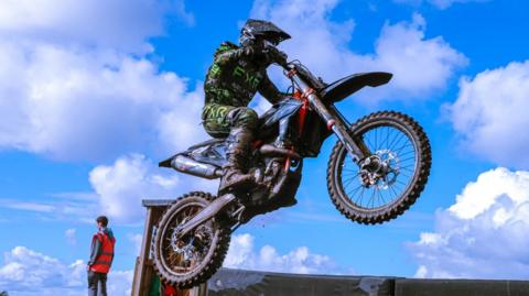 A dynamic moment as a person, riding a dirt bike, is caught mid-air during a jump. The rider is equipped with protective gear, including a helmet and clothing featuring green and black patterns. In the background, there's another individual wearing an orange vest, possibly an official, standing near a small structure. 