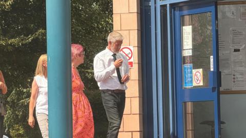 A man in a white shirt, black tie and dark jeans walks into a court building with three women. He has silver hair and a beard.