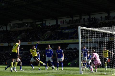 Footballer Myles Peart-Harris strikes a football for Oxford United in a crowded penalty area during a Championship match at home to Watford. The Oxford players are wearing a yellow kit with dark blue shorts and trim details, while Watford are wearing blue shirts and socks with white shorts. A goalkeeper in a predominantly pink kit is visible on the goal line, as is a packed stand full of fans at the Kassam Stadium