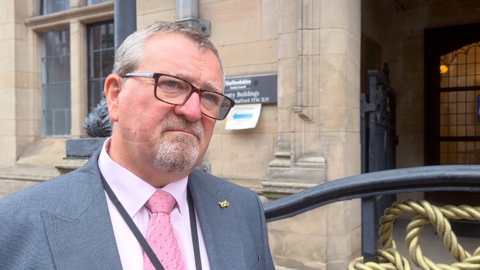 A head and shoulders shot of Peter Mason, who has grey hair and a neat grey beard. He has glasses, is wearing a white shirt with a pink tie and a grey-blue suit. He is standing in front of a council building