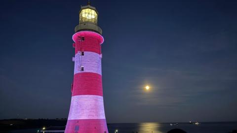 Plymouth lighthouse lit up at night. In the background is the sea, with the moon reflecting on the water. The lighthouse is red and white and there is a light on at the top.