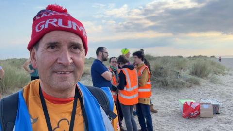 Trevor Rawson standing on the beach. He has on a blue high visibility vest, an orange T-shirt and is wearing a red hat. He is smiling. A group of volunteers are in the background. 