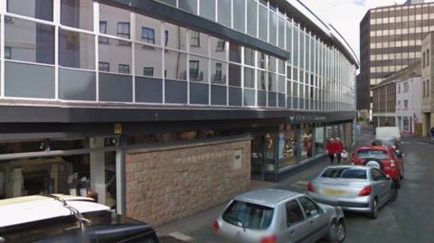 A picture of Dumaresq Street. There are a number of cars parked to the right of the image. There are two lorries parked in the distance.