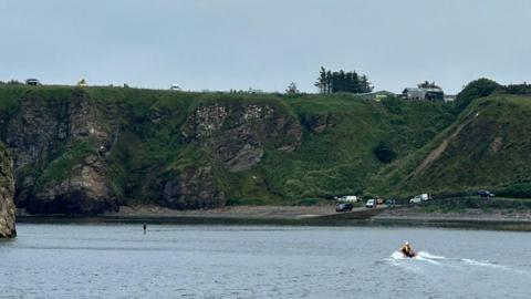 A figure can be seen standing on an almost submerged rock near a harbour. There is a small rescue boat going to his aid.  In the background there is a stony beach and some cliffs. 