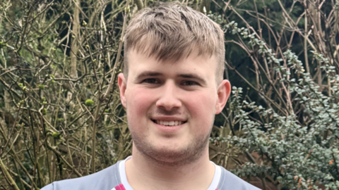 Samuel is standing outside by a hedge in a pink and grey sports top. He is smiling at the camera and has short brown hair a short beard.