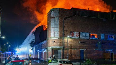 Massive orange flames above the former cinema and fire brigade are spraying water at it. A red car and a white car are on the road next to it. 