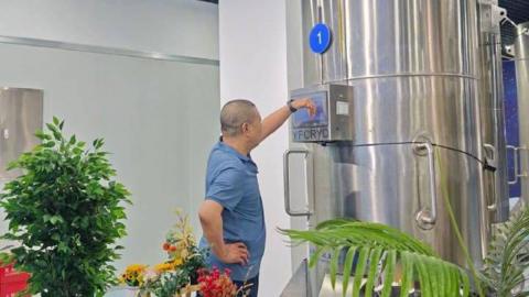 A man in a blue t-shirt stands looking at a large metallic container which has the letters YFCYRO on it. Around him are several green plants.