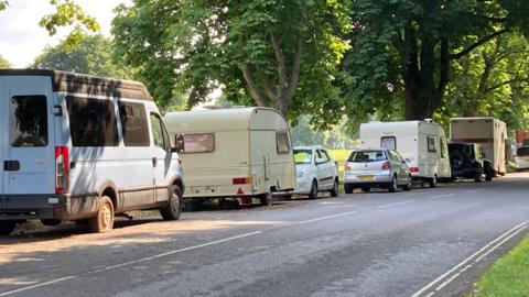 A row of caravans and vans on the side of the road with a few cars in between, they are parked next to large grassy open spaces with large trees.