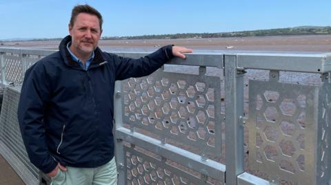 A man called Stuart Greenslade stands next to a fence on a train station platform. He is wearing a navy blue coat. He has brown trimmed hair and a trimmed grey beard. The River Exe is behind the railings.