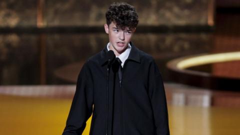 Young actor Owen Cooper wears a black jacket, black tie and white shirt and stands on stage with microphone in front of him.