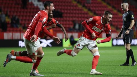three charlton players wearing red shirts, white shorts and red socks run together with mouths agape and joyful expressions on their faces.