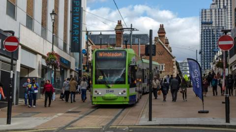 A green tram in a shopping street in Croydon. There are shoppers on either side of the tramway