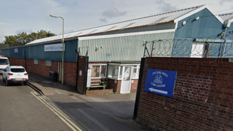 A corrugated metal warehouse at Jacobs Well Care Centre. The entrance gate is between brick walls topped with coiled, barbed wire.