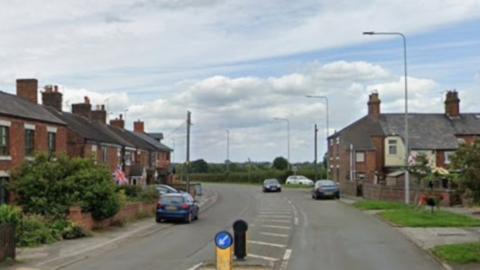 A single carriageway road with traditional properties on either side and a couple of cars parked up on the edge of the road. Beyond the houses, hedging sits along the edge of fields.