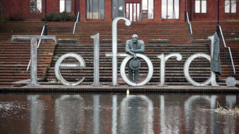 A large metal sign behind a small pond which reads Telford with red brick steps behind