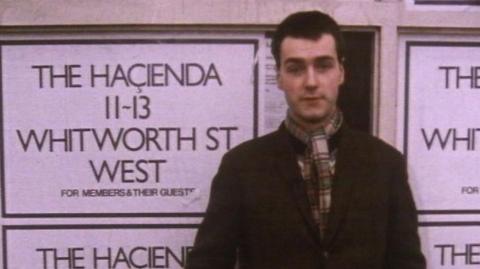 Fraser Diagram stood  in front of posters for the Hacienda, wearing a scarf and a dark coat, looking directly down the lens of the camera.  The text on the poster behind reads: THE HACIENDA, 11-13, WHITWORTH ST, WEST, FOR MEMBERS & THEIR GUESTS