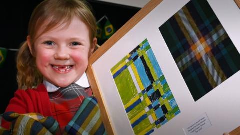A six-year-old girl holds up a framed sample of tartan in her arms. She is smiling at the camera.
