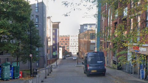 A narrow urban street lined with mid-rise buildings on both sides, featuring a mix of brick and modern facades. On the left, there are stacked recycling bins and a large tree with green foliage. On the right, a convenience store and a parked black van.
