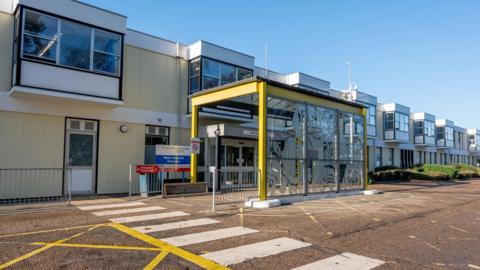 The exterior of The Queen Elizabeth Hospital. It is a modern two-storey building painted in blue and yellow. A pedestrian crossing is painted on the road in the foreground.