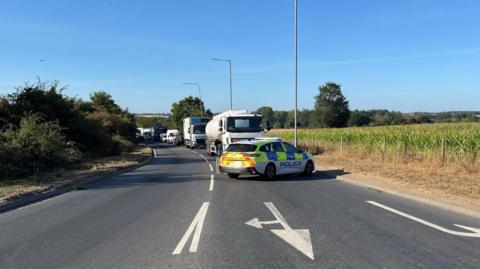 A police car is parked at an angle across a road, with traffic queued behind it. The other side of the road is empty.