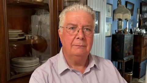 A man with grey hair, which is combed back, is sitting in a chair. He is wearing glasses and a pink, open collared shirt with pinstripes. Behind him is a collection of antique furniture and household items including a lamp and photographs. There is also a white cottage style door separating two rooms.