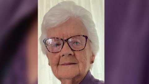 A woman in her late 80s smiles softly. She has short white hair, is wearing pattern rimmed glasses, flower stud earrings and a purple blouse. There is a white curtain behind her.