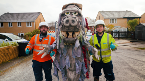 Two men in white hard hats, one wearing an orange hi-vis top and the other wearing a yellow hi-vis vest, looking at the camera, holding their thumbs up. They are stood either side of a person wearing a fatberg monster costume.
They are stood on a road of a residential street.