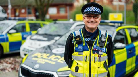 A police officer standing in a high-viz gilet in front of a yellow and blue marked police car, wearing a checked police hat.