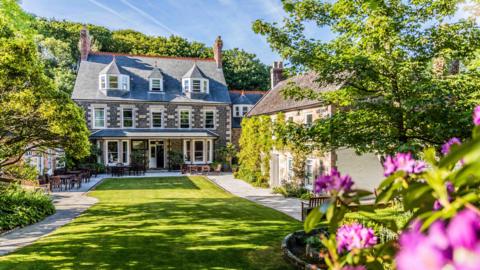 The image shows a view of the Stocks Hotel HQ garden. There is a manor house in the distance, with a long grass area and flowers surrounding it.