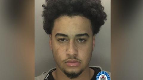 A mugshot of a man who has a short beard and thin moustache. He has brown eyes and an afro. He is staring into the camera and is wearing a grey hoodie over a black top. 