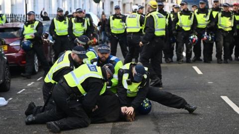 Police officers detaining a man in the road, holding him to the ground, while other officers stand nearby in a line
