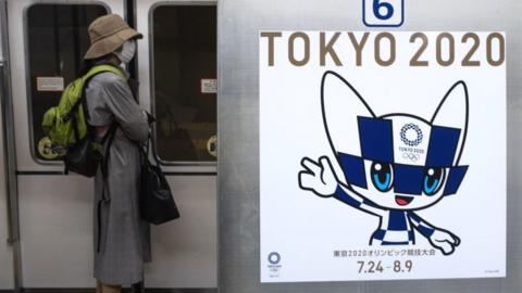 A passenger wearing a face mask stands next to a poster of Tokyo 2020 Olympic mascot Miraitowa on a train in Tokyo on April 20, 2020.