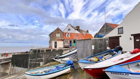 Sever small rowing boats sit on a deck with buildings behind. Beyond the sea the blue sky if streaked with white cloud