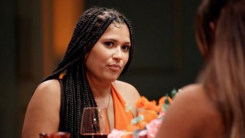 Tasha on Married At First Sight UK looking at a fellow contestant. She is wearing an orange outfit, sitting at a table where a wine glass and orange and pink flowers are visible. It's an over the shoulder shot from the opposite side, with the background blurred and dark.