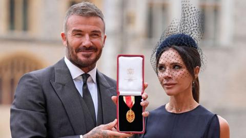 Sir David Beckham, on the left, wearing a grey suit and holding a box containing a red and gold medal, next to Victoria Beckham, who is looking at him smiling, wearing a navy outfit and a small hat with a veil