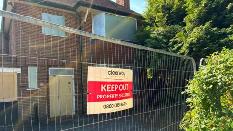 Boarded up house behind security fencing, with a Keep Out sign on it