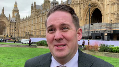 Close up of Sam Rushworth smiling at looking at the camera. He is standing in front of Big Ben and Parliament which has some scaffolding on. It is a grey day and he is standing on some grass near the the Gothic Revival structures.