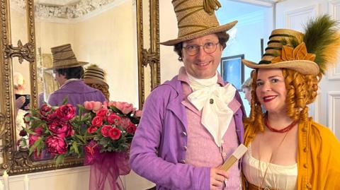 A man in a purple suit with a wicker top hat and he holds a fan. Next to him is a lady in yellow dress with her hair in ringlets, red lipstick and a similar wicker hat